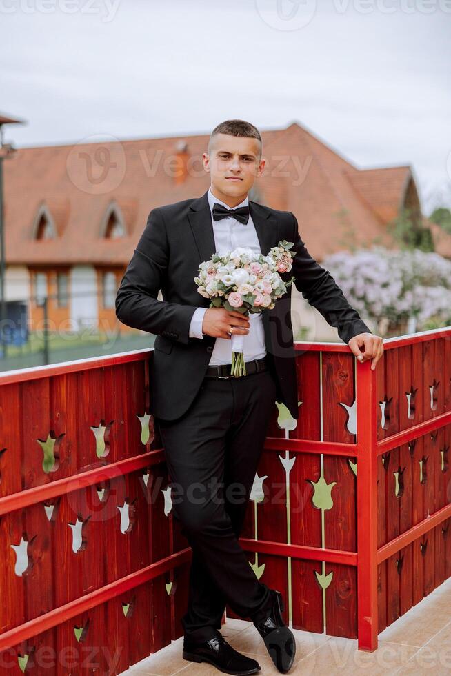 The groom in a black suit holds a bouquet, poses leaning on a red railing. Wedding portrait. photo