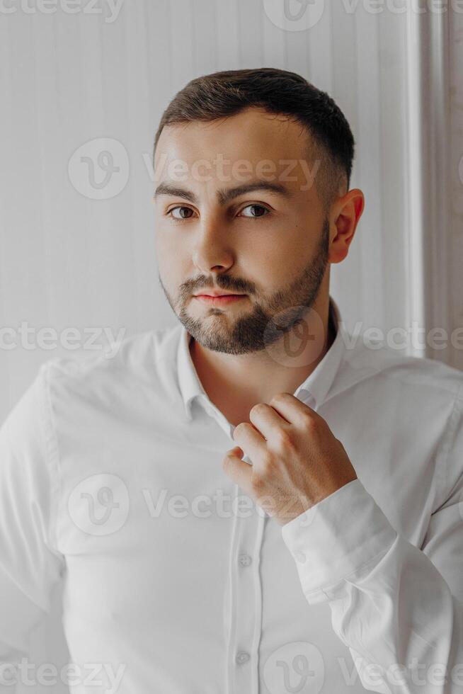 Handsome man putting on a shirt by the window in a hotel room in the morning. Preparation for an event or a new working day. New opportunities, acquaintances. Close-up portrait of an adult man photo