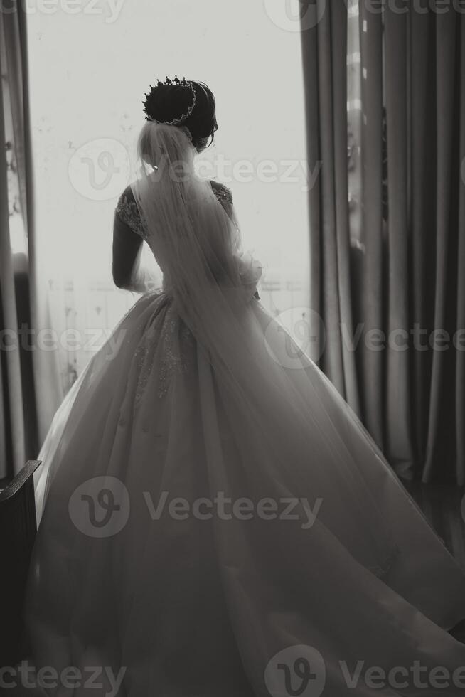 un lujoso novia con un hermosa peinado y un elegante vestido, espalda vista, es consiguiendo Listo para el Boda ceremonia en el Mañana. Mañana foto de el novia a hogar o en un hotel habitación.
