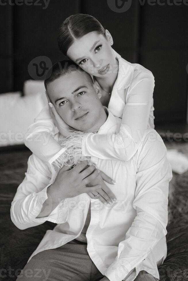 joyful young bride in white pajamas hugs the shoulders of handsome groom in white unbuttoned shirt while sitting together in modern hotel room before wedding ceremony. Black and white photo