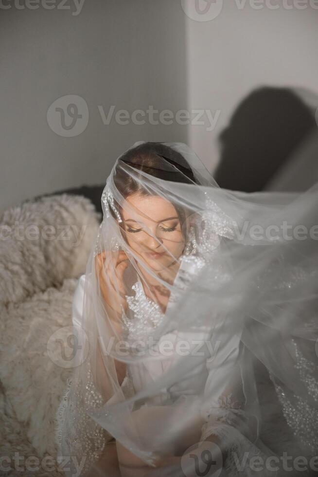 Nice cute girl in a boudoir robe. Happy beautiful bride under veil closeup. Fashion, beauty, style. Morning of the bride. Beautiful bride with stylish make up and hair style. photo