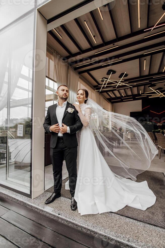 Cute newlyweds are gently hugging and smiling near the restaurant. Portrait of the bride and groom in a lace dress. photo