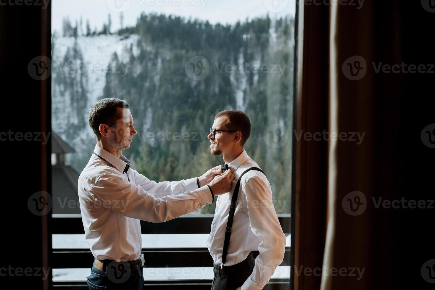 un respetuoso padre ayuda su maduro hijo poner en un arco Corbata antes de el boda. disparo en el hotel en contra el antecedentes de nevadas montañas foto