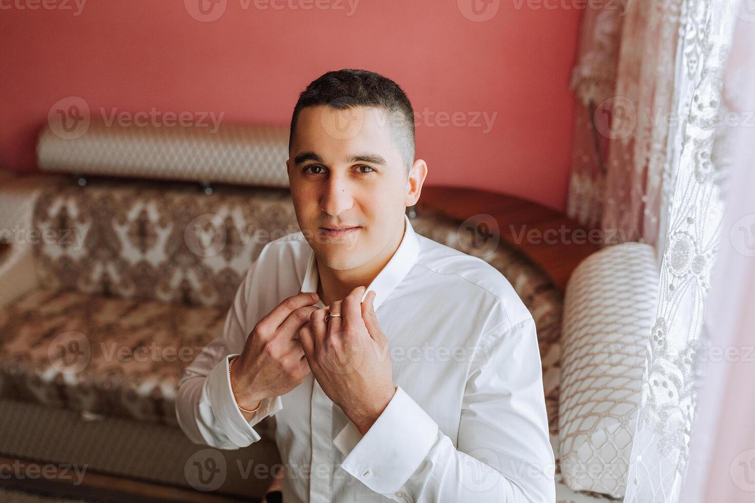 Handsome business man buttoning shirt collar at home. The groom is preparing for the wedding. Close-up portrait. photo