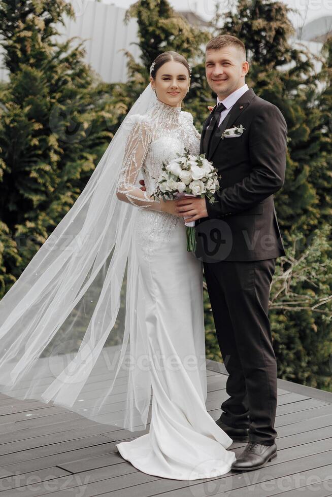 retrato. un novia en un elegante largo vestir y velo y un novio en un traje actitud mientras en pie cerca arboles Boda en naturaleza foto