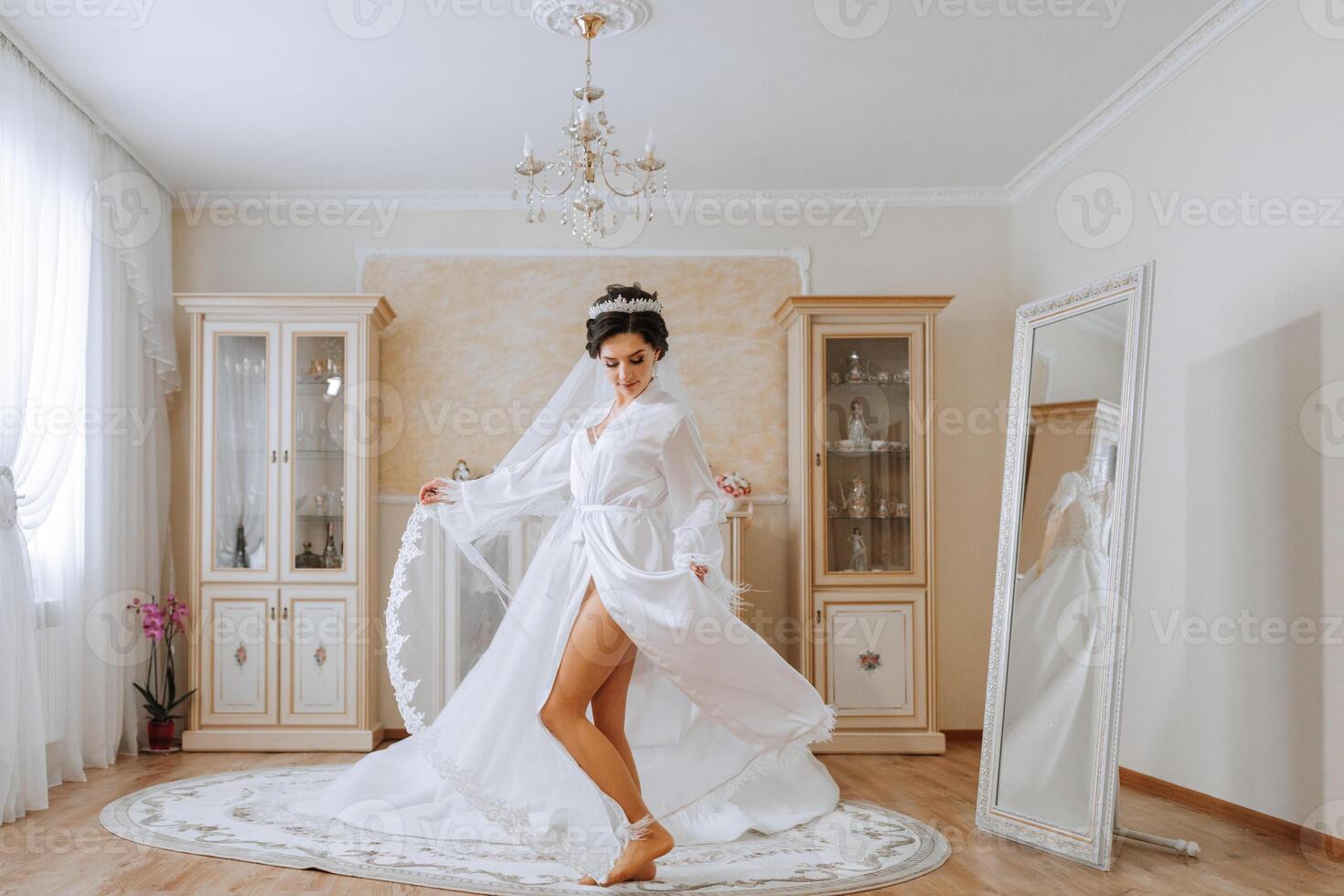 Morning of the bride before the wedding. A beautiful young woman with a veil and a beautiful hairstyle in a white robe near her wedding dress on a mannequin. Professional makeup. Last preparations. photo