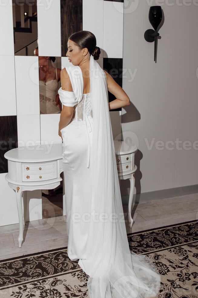 Back view of a bride in a luxurious elegant wedding dress with a long veil standing near a mirror in a bright studio. photo