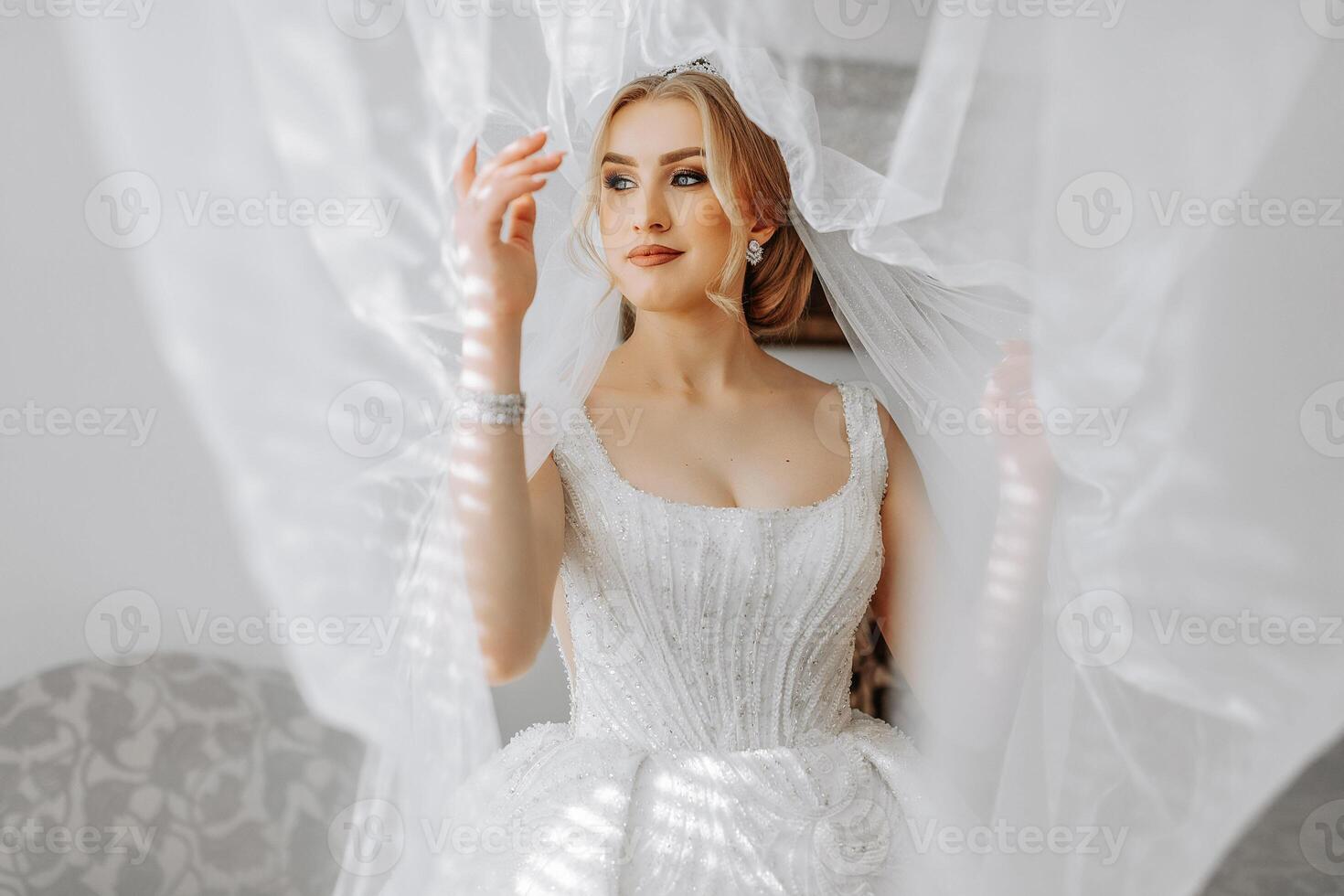 el novia en un blanco vestido, cubierto con un velo, poses en bueno Encendiendo en su habitación. Mañana de el novia. un hermosa tiara foto