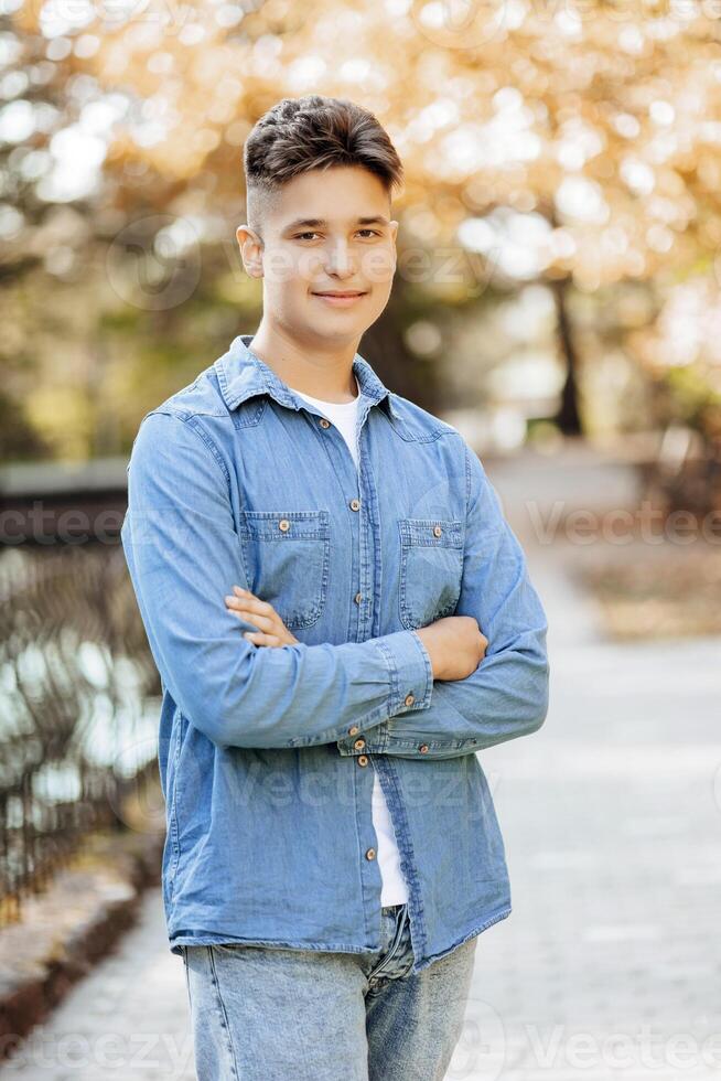 Vertical close-up portrait of a teenager in casual clothes. Happy smiling teenager in autumn park in sunlight. A beautiful child looks at the camera in nature. photo
