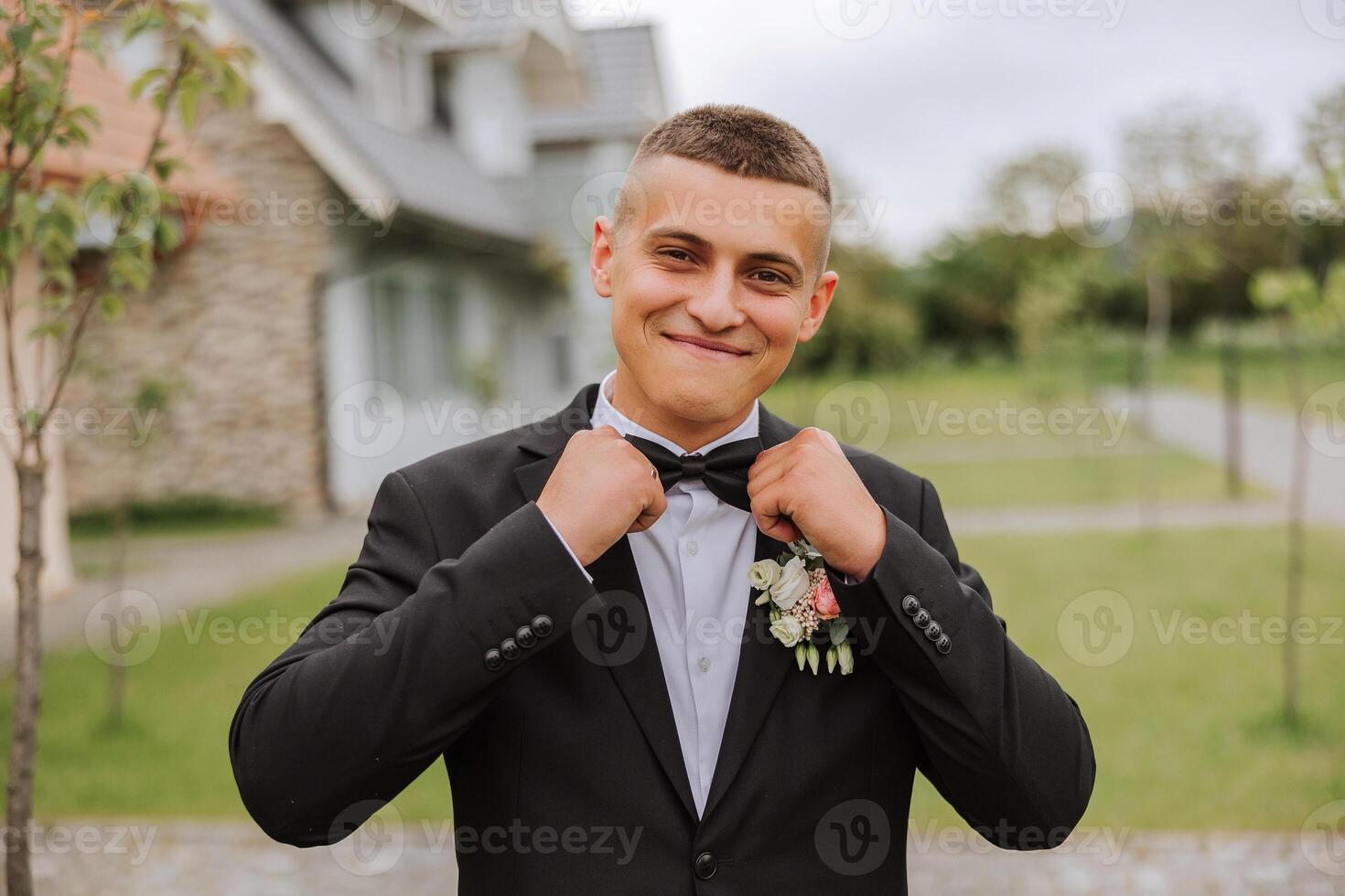 The groom in a black suit adjusts his bow tie, poses against the background of a green tree. Wedding portrait. photo