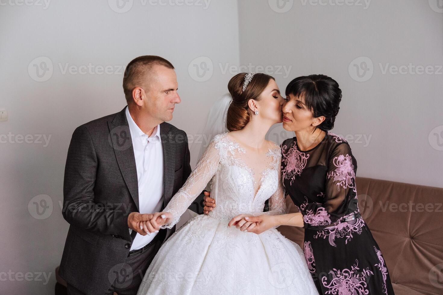 Happy parents hold the hands of the bride who is getting married and support her, wearing a wedding dress and a long veil. photo