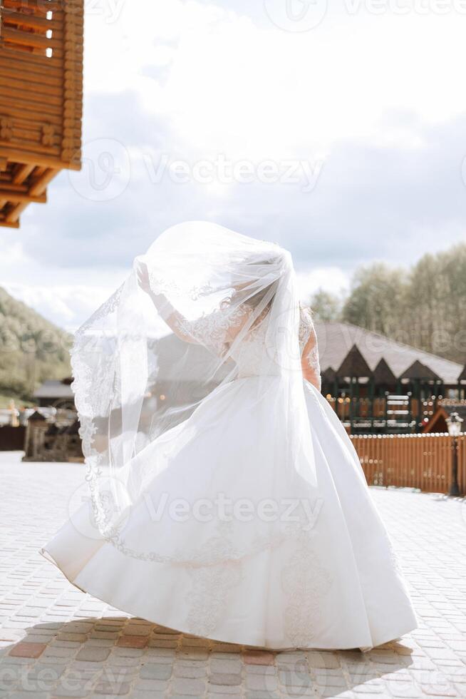 A curly bride twirls her long veil against a backdrop of mountains and wooden houses. Magnificent dress with long sleeves, open bust. Summer wedding photo