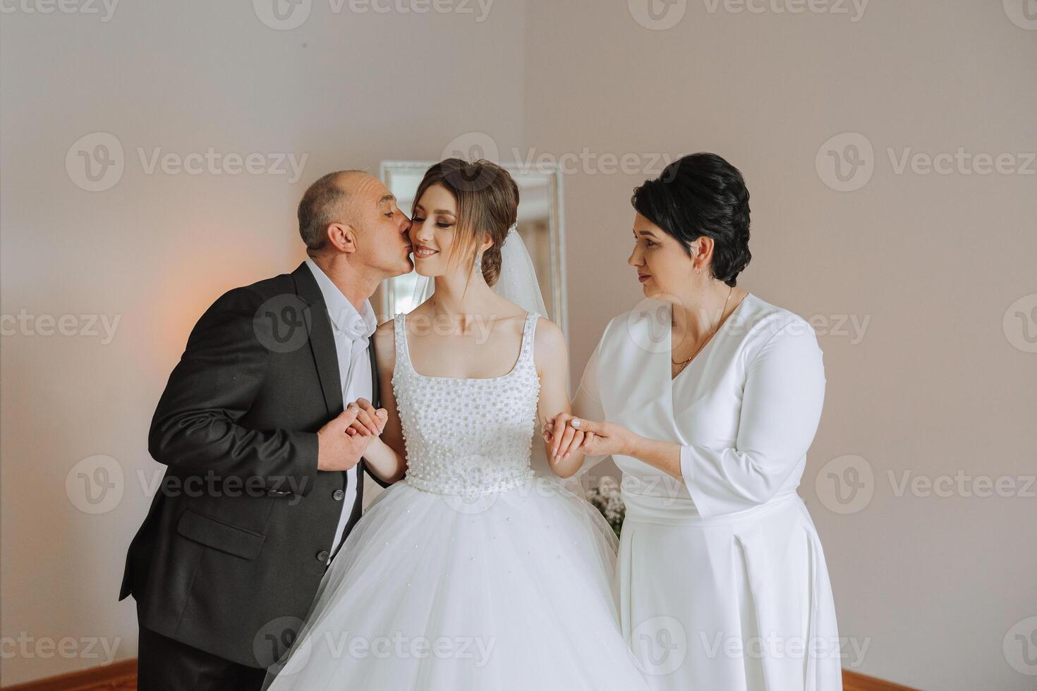 Happy parents hold the hands of the bride who is getting married and support her, wearing a wedding dress and a long veil. photo