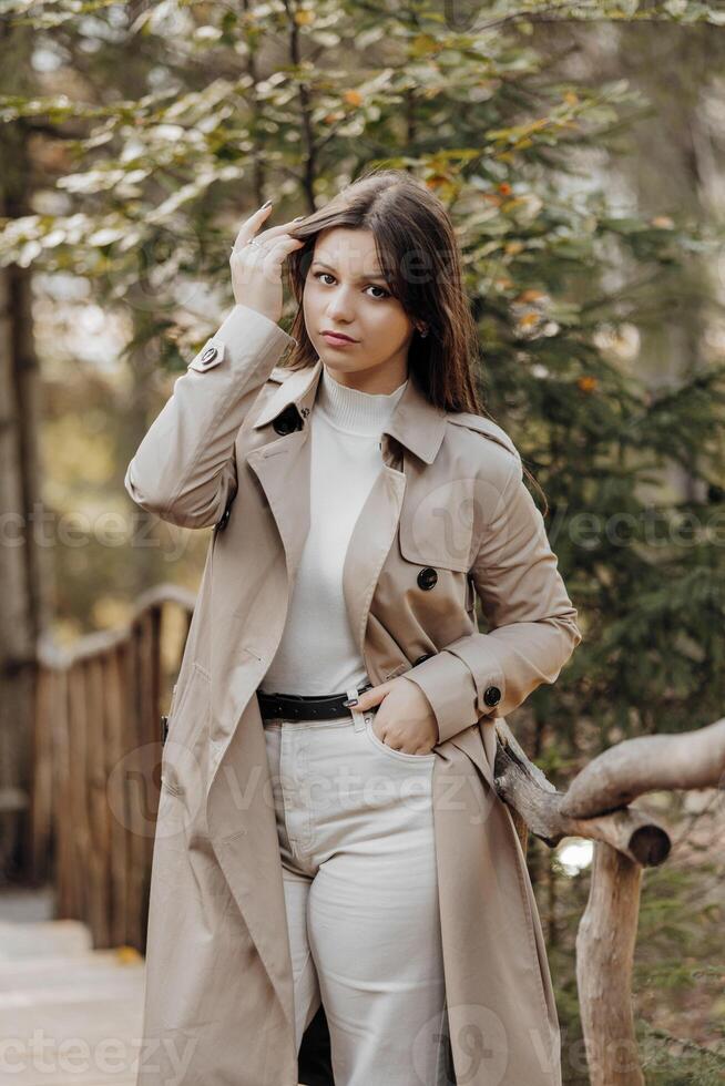 Vertical photo. Beautiful carefree young teenage girl in brown coat and white jeans and white sweater. Portrait of a beautiful girl in a brown raincoat against the background of the forest and nature. photo