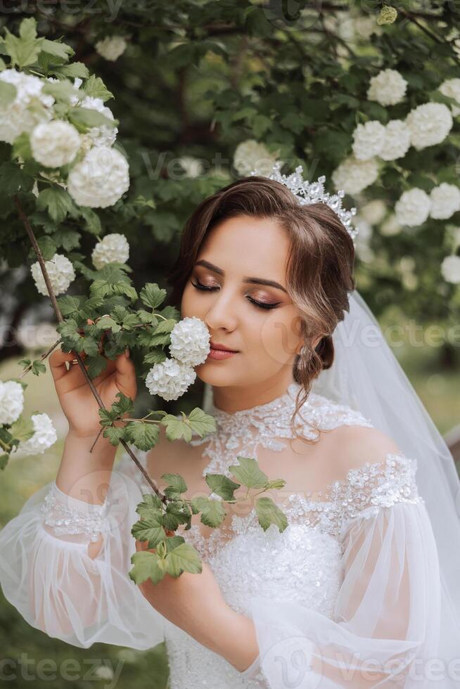 Curly brunette bride in a lush veil and long-sleeved dress poses next to a white lilac. Beautiful hair and makeup. Spring wedding photo