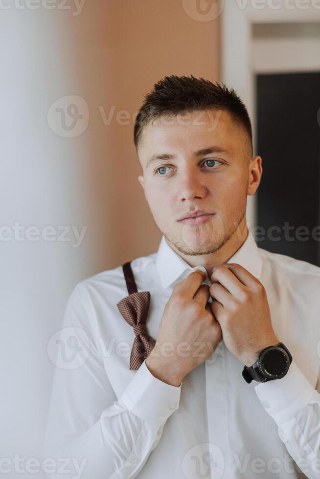 The groom buttons his shirt by the window. man in a shirt. Male portrait. Photoshoot at the window. Groom's collection. Wedding day. Handsome man groom buttoning his shirt in a hotel room. photo