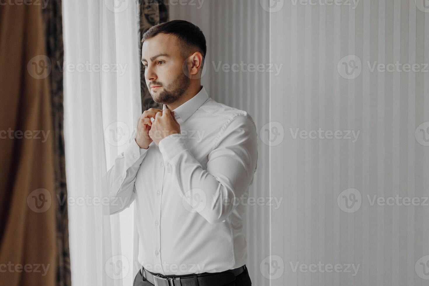 Handsome man putting on a shirt by the window in a hotel room in the morning. Preparation for an event or a new working day. New opportunities, acquaintances, wedding day or preparing for an interview photo