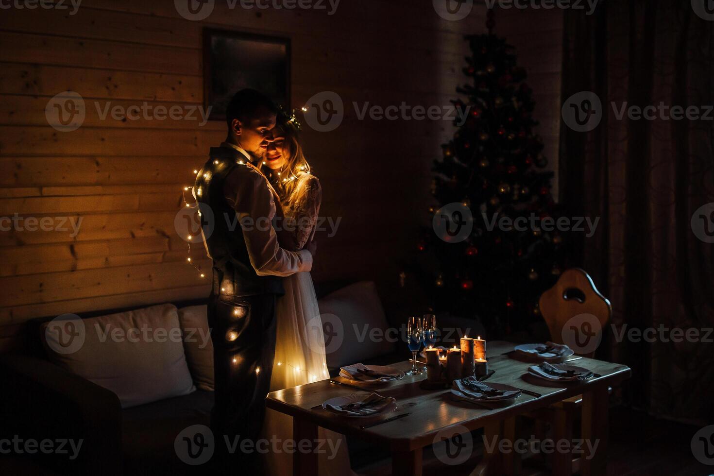 a happy bride and groom embrace in a dark room illuminated by defocused lights of a garland photo