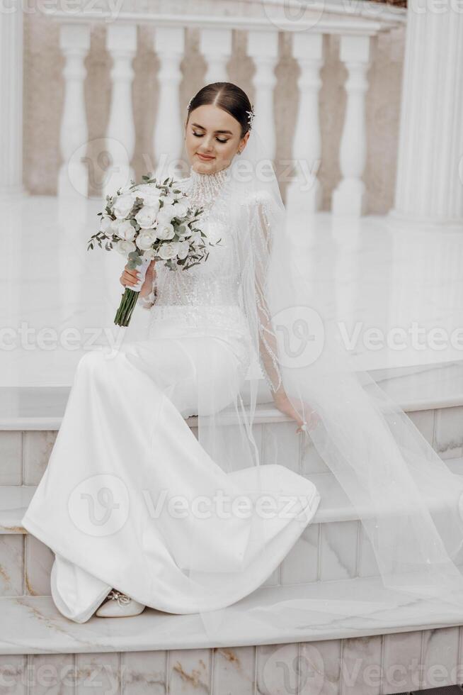 The bride in a long dress sits near white Roman-style columns. Beautiful hair and makeup. An exquisite wedding photo