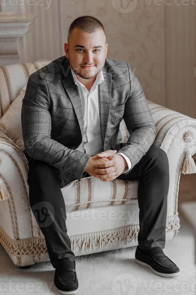 a handsome and positive man is sitting on an expensive chair. Smiling at the camera, wearing smart shoes, trousers, a jacket and a white shirt photo