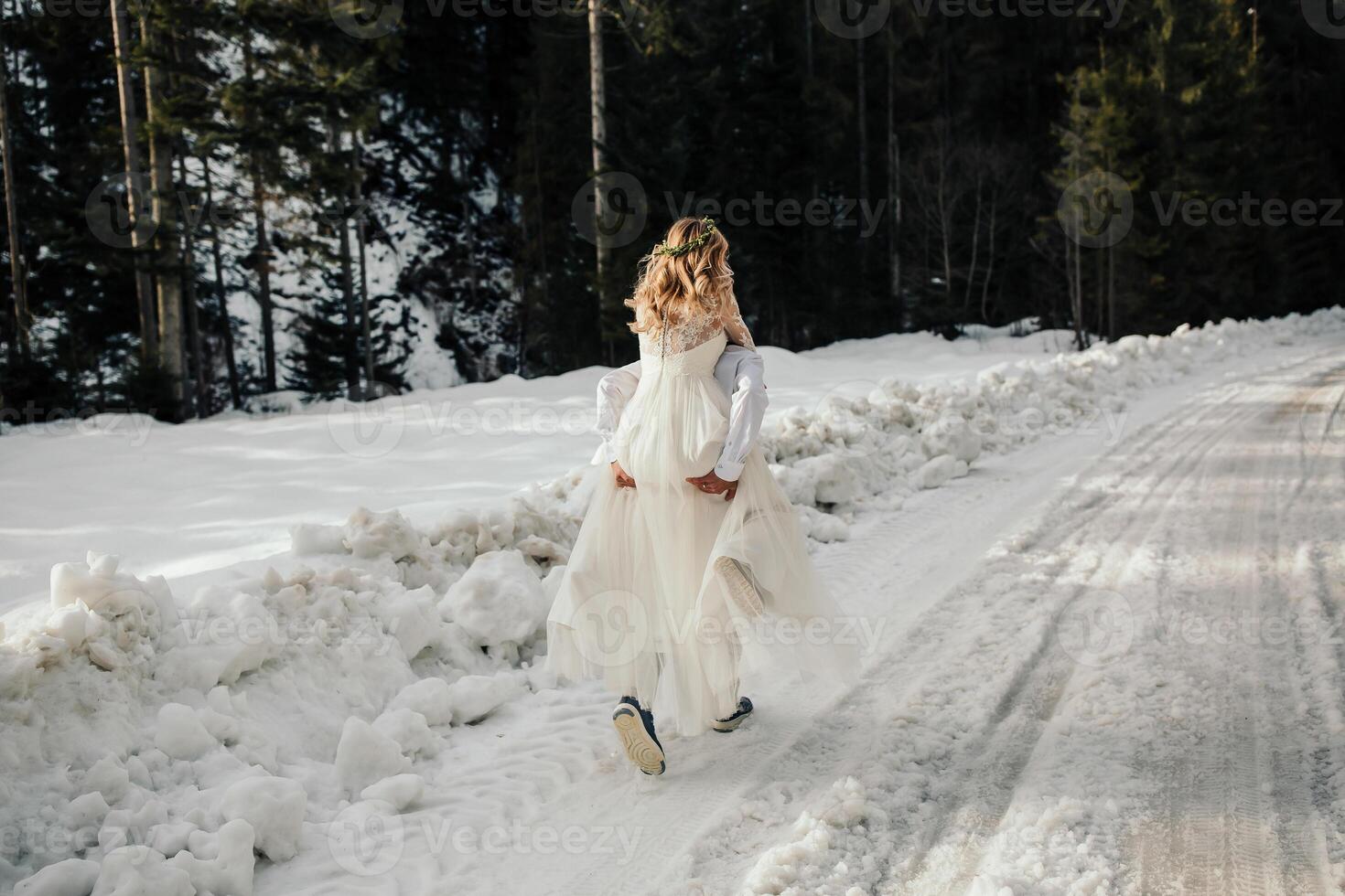 The bride and groom are running along a snowy road against the background of a pine forest and beautiful contrast sunlight. Side view. Winter wedding. Place for logo. photo