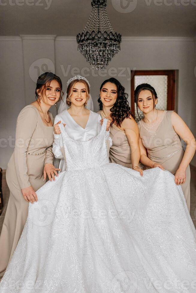 The bride in a white robe and her friends in cream dresses pose near the bride's dress. Morning of the bride photo