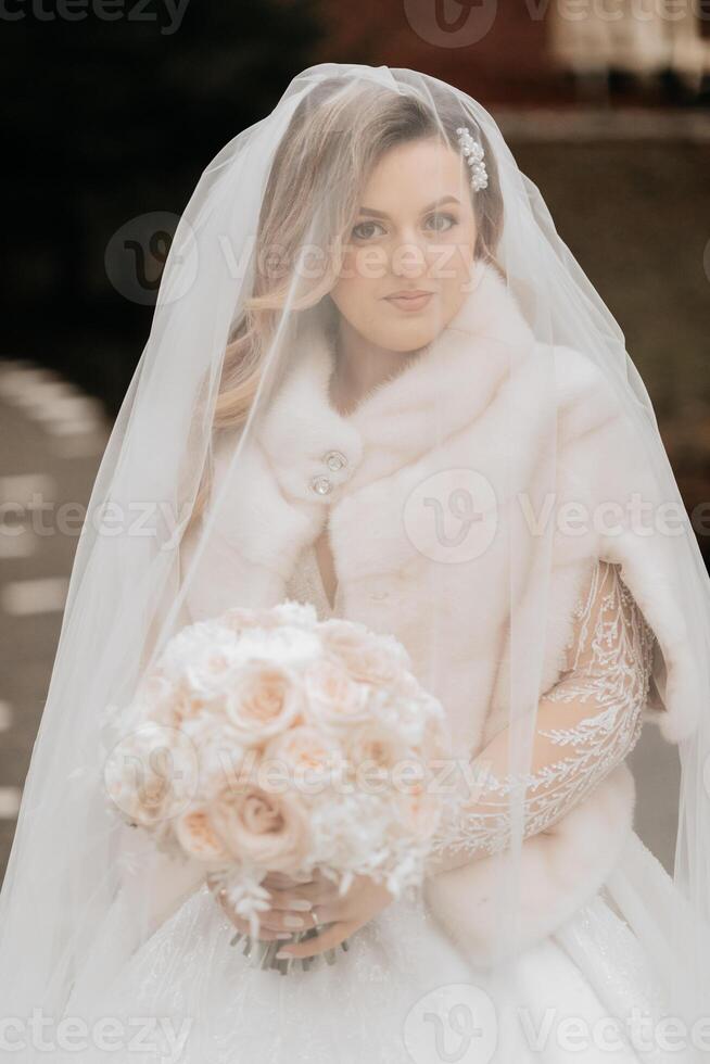 Portrait of a beautiful bride with a wedding bouquet of flowers, attractive woman in a wedding dress with a long veil. Happy bride woman. Bride with wedding makeup and hairdo. Winter wedding photo