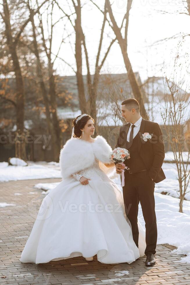 Winter wedding. Happy couple walking in wedding clothes hugging and smiling in a winter park covered with snow on their wedding day. Winter love story of a beautiful couple in snowy winter weather photo