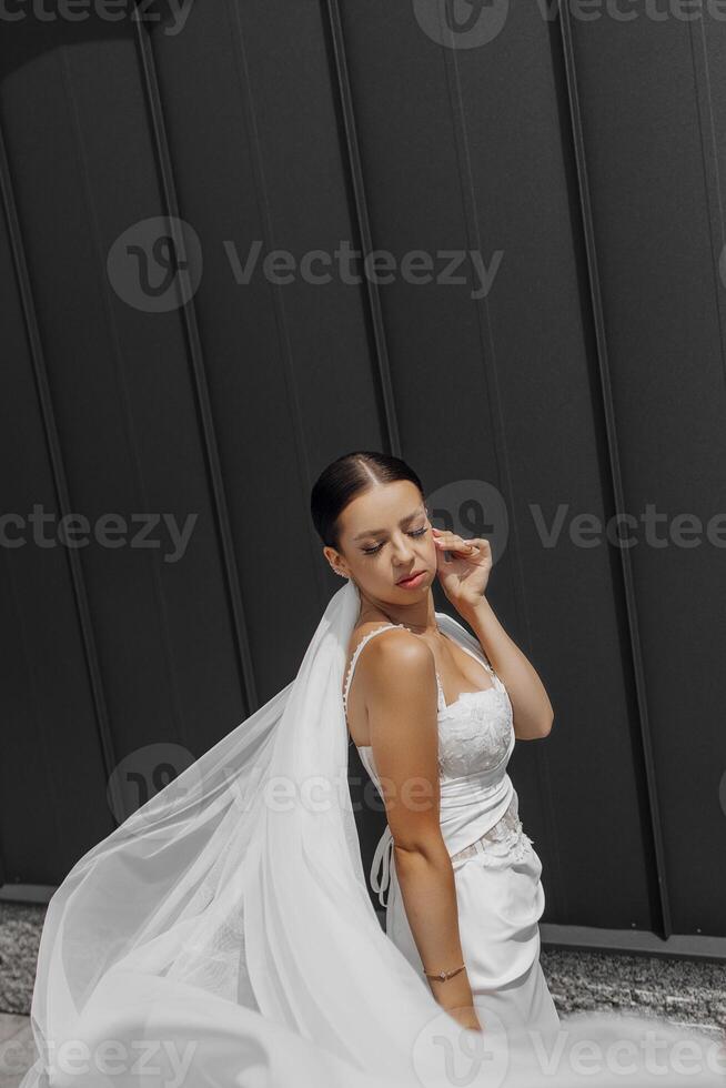 hermosa joven novia niña vistiendo un blanco vestir con un largo velo y posando cerca un gris pared en un moderno ciudad. foto