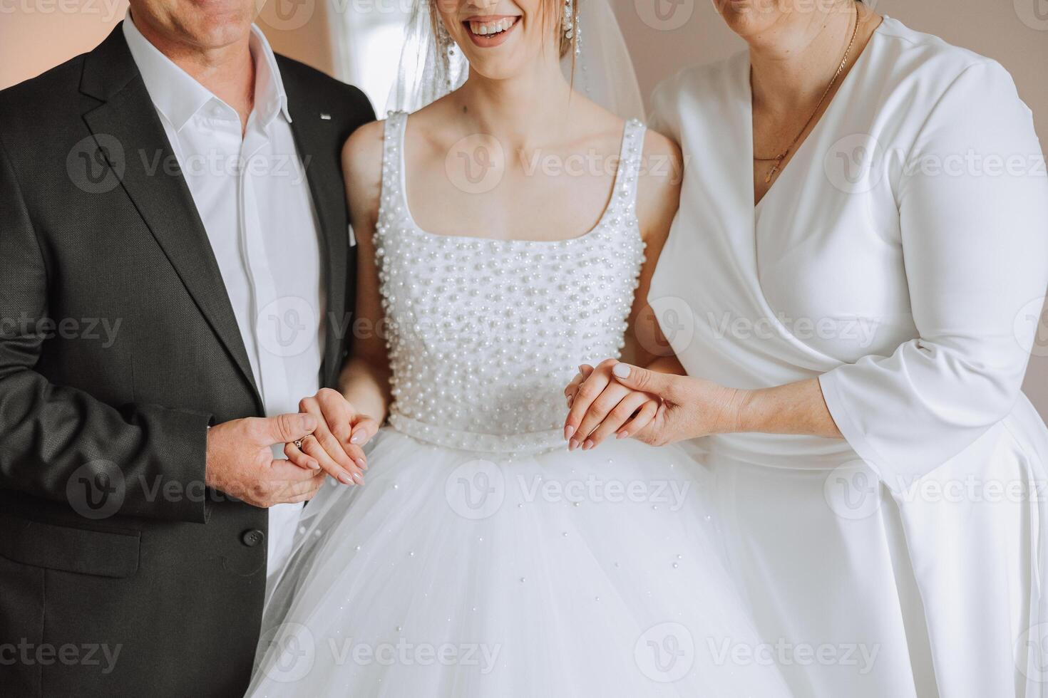 Happy parents hold the hands of the bride who is getting married and support her, wearing a wedding dress and a long veil. photo