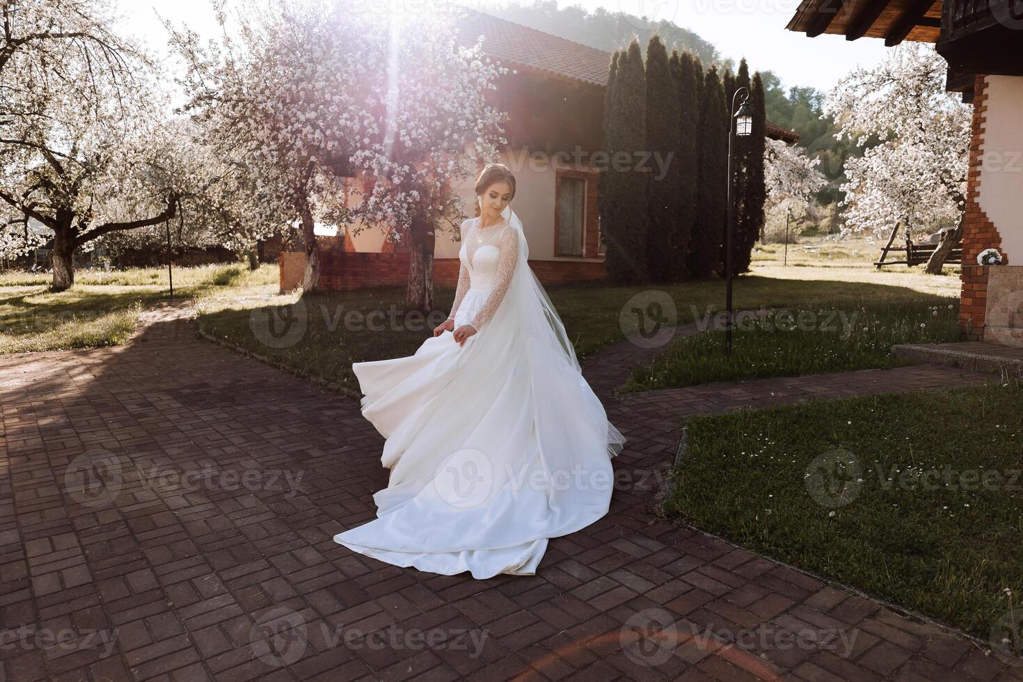 The bride has her shoulders turned to the camera, walking along a stone path. A long train. Magnificent dress with long sleeves, open bust. Spring wedding photo