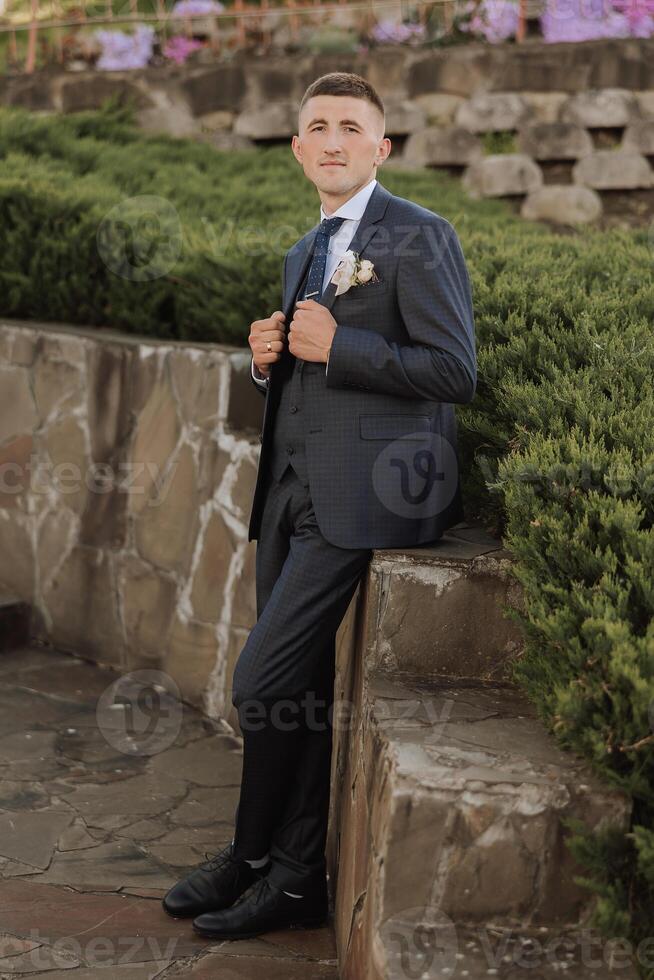 The groom in a black suit adjusts his jacket, poses against the background of a green tree. Wedding portrait. photo