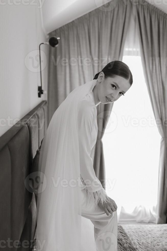 A beautiful and confident bride standing on a bed in white pajamas with a long veil in a hotel room poses for a photographer. Wedding concept. Morning of the bride. Preparation for the wedding. photo