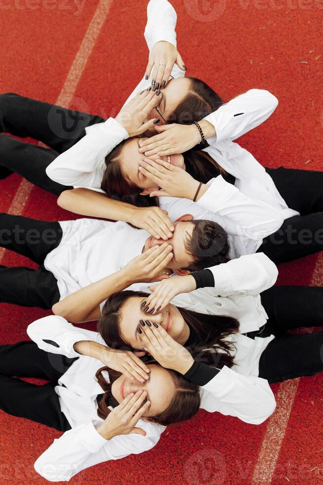 Happy friends lying together in a circle looking at the camera, holding hands and smiling while lying on a red wall... Friendship, lifestyle, togetherness, business and teamwork concepts. photo