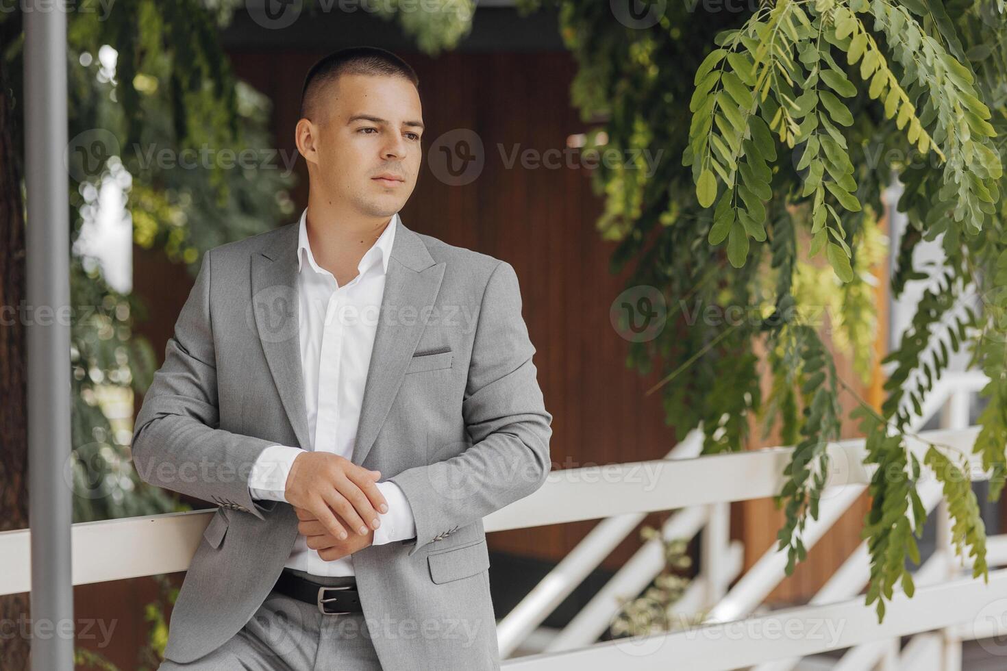 Handsome successful young man wearing gray suit, white shirt, sunglasses and posing near green trees. photo
