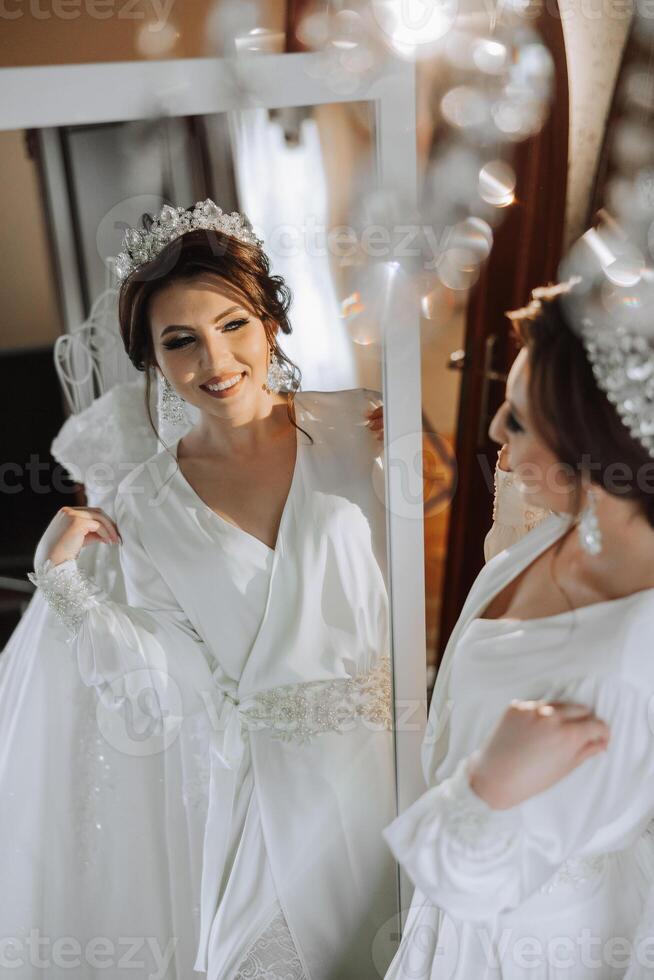 portrait of a luxurious bride in boudoir style near a mirror with reflection. Preparing the bride for the wedding in the morning in a white robe. photo