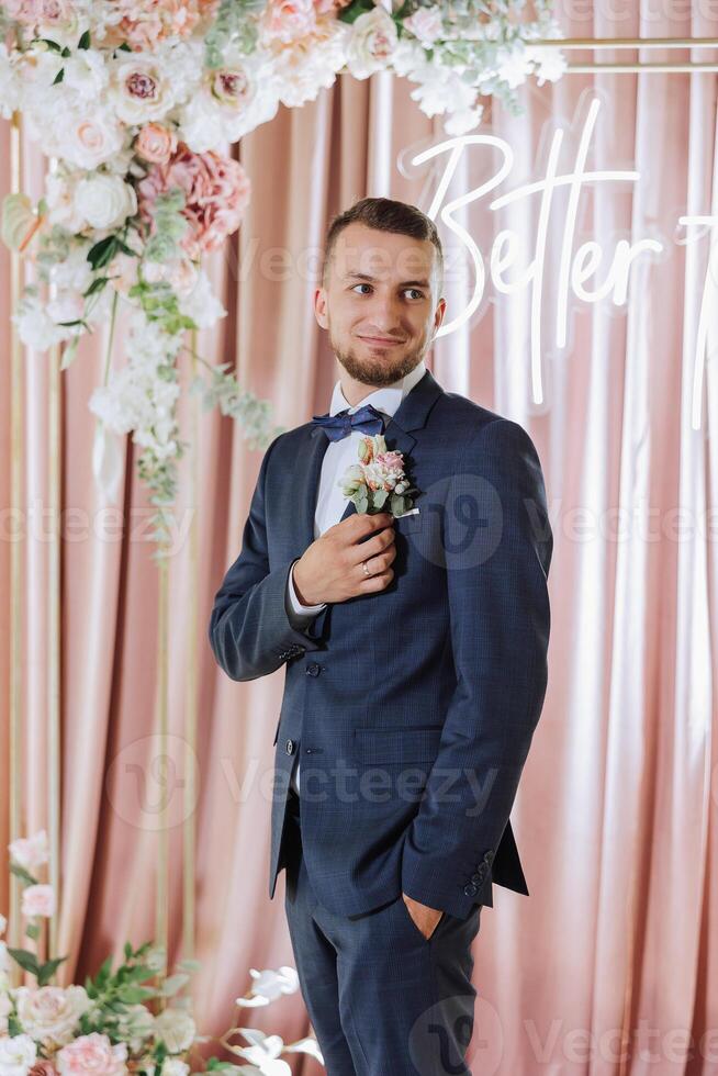 The groom in a black suit adjusts his boutonniere, poses against the background of a pink photo booth. Wedding portrait.