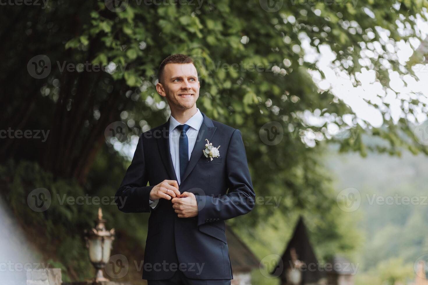 The groom in a black suit adjusts his jacket, poses against the background of a green tree. Wedding portrait. photo