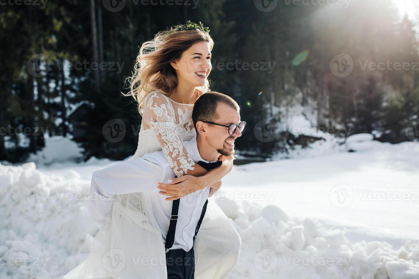 The bride and groom are running along a snowy road against the background of a pine forest and beautiful contrast sunlight. Side view. Winter wedding. Place for logo. photo