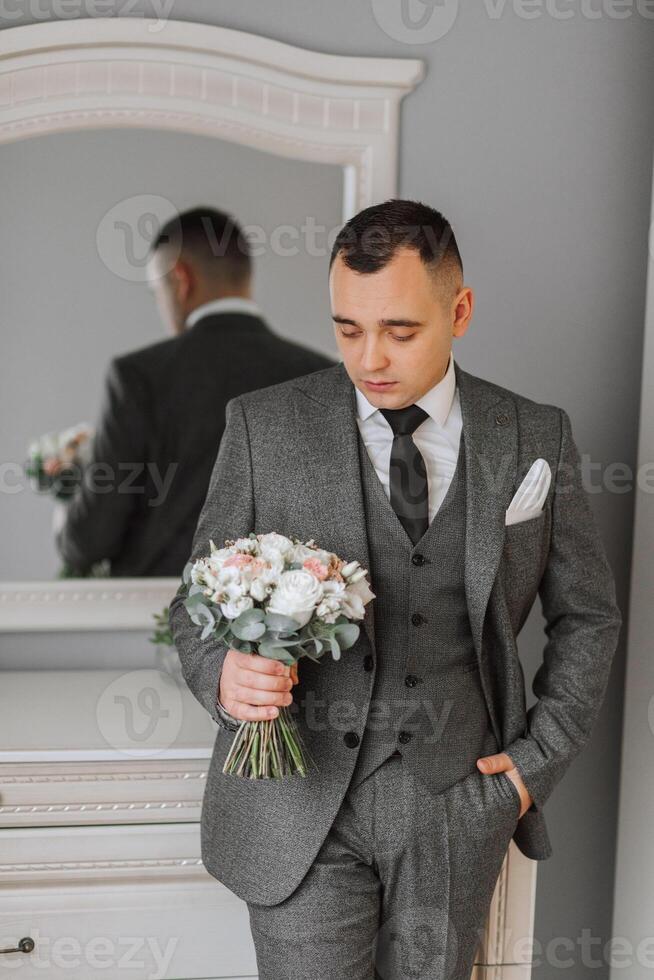 A man in a gray suit is holding a bouquet, posing in his room. Portrait of the groom. Fashion and style. Business photo