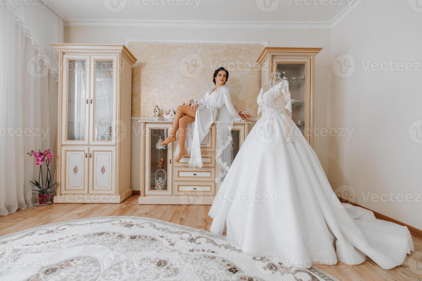 Mañana de el novia antes de el boda. un hermosa joven mujer con un velo y un hermosa peinado en un blanco túnica cerca su Boda vestir en un maniquí. profesional constituir. último preparativos. foto