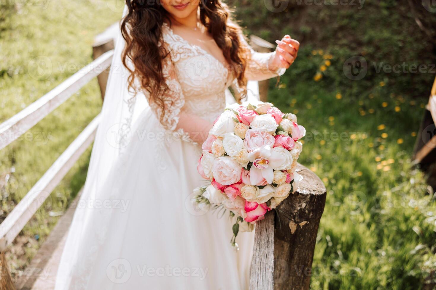 Curly brunette bride poses with a bouquet, near a wooden railing. Magnificent dress with long sleeves, open bust. Spring wedding photo