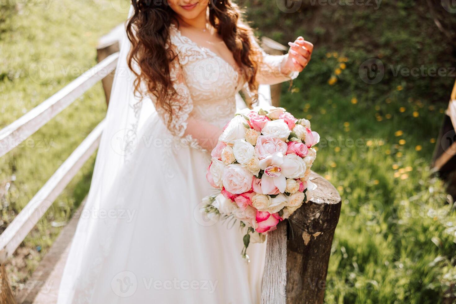 Rizado morena novia poses con un ramo, cerca un de madera barandilla. magnífico vestir con largo mangas, abierto busto. primavera Boda foto