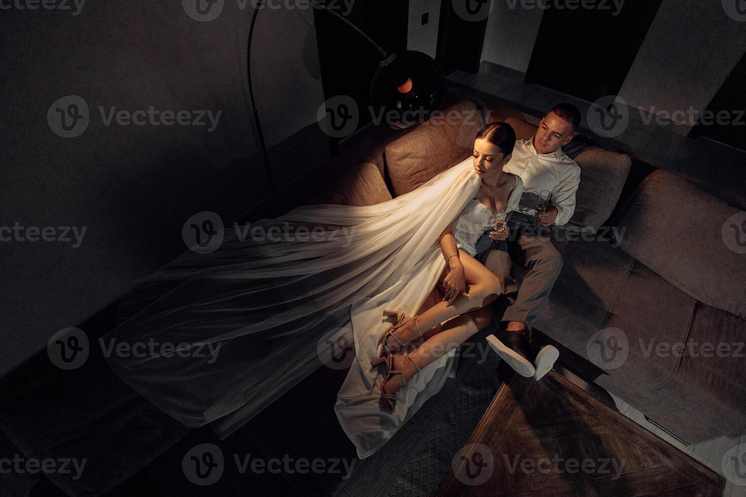 retrato de el recién casados en el hotel acostado en el sofá con lentes de champán. el novio suavemente toques el novia. después el Boda ceremonia. contento novia y novio celebrando foto