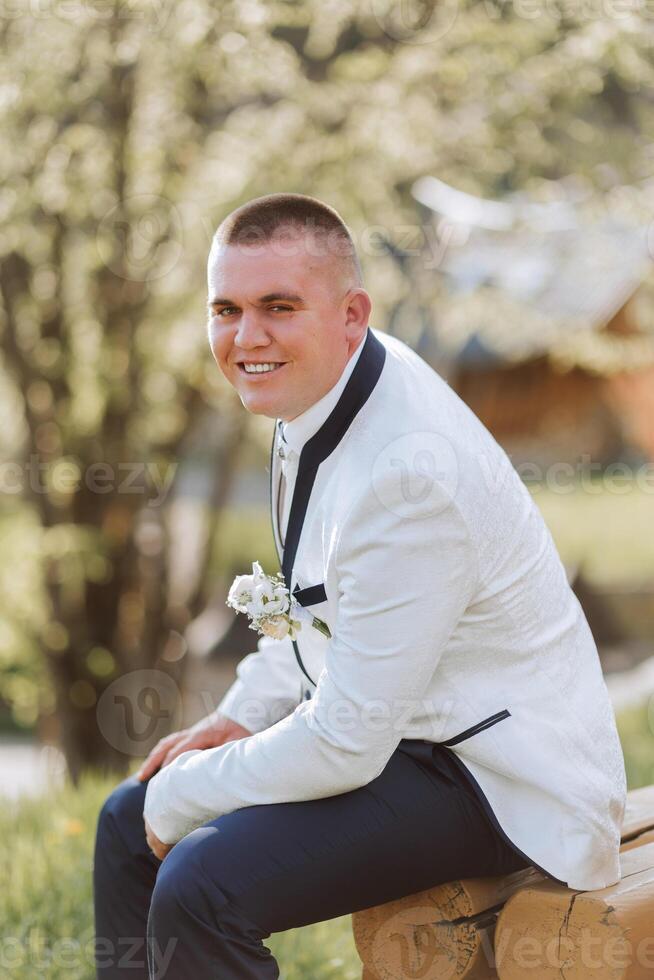 The groom in a white jacket and black trousers poses while sitting on a wooden chair. Wedding portrait. photo