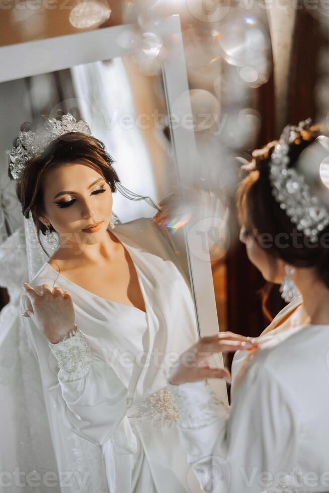 portrait of a luxurious bride in boudoir style near a mirror with reflection. Preparing the bride for the wedding in the morning in a white robe. photo