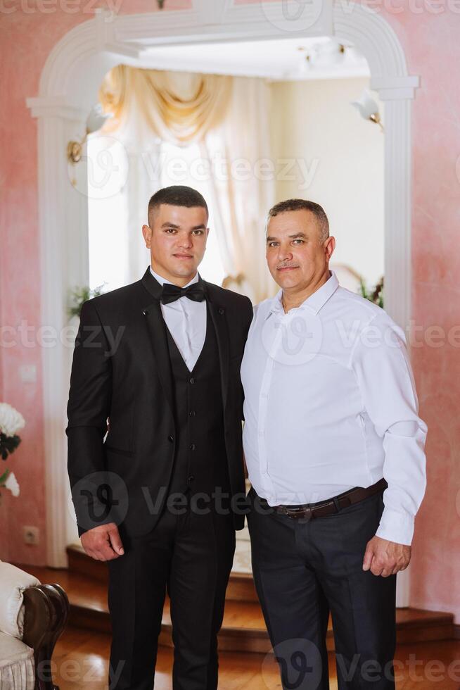 un padre abrazos su maduro hijo y ayuda él preparar para el Boda ceremonia. calentar y sincero relaciones Entre un padre y un adulto hijo. un emocional momento a un Boda foto