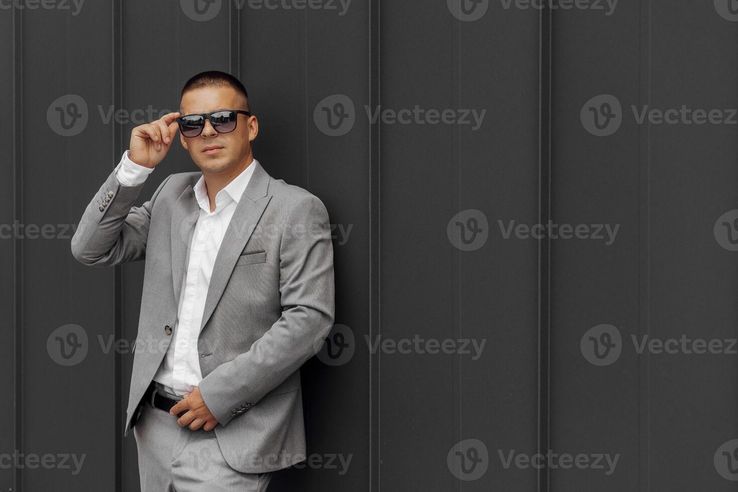 hermoso exitoso joven hombre vistiendo gris traje, blanco camisa, Gafas de sol y posando en contra gris pared en moderno ciudad. foto