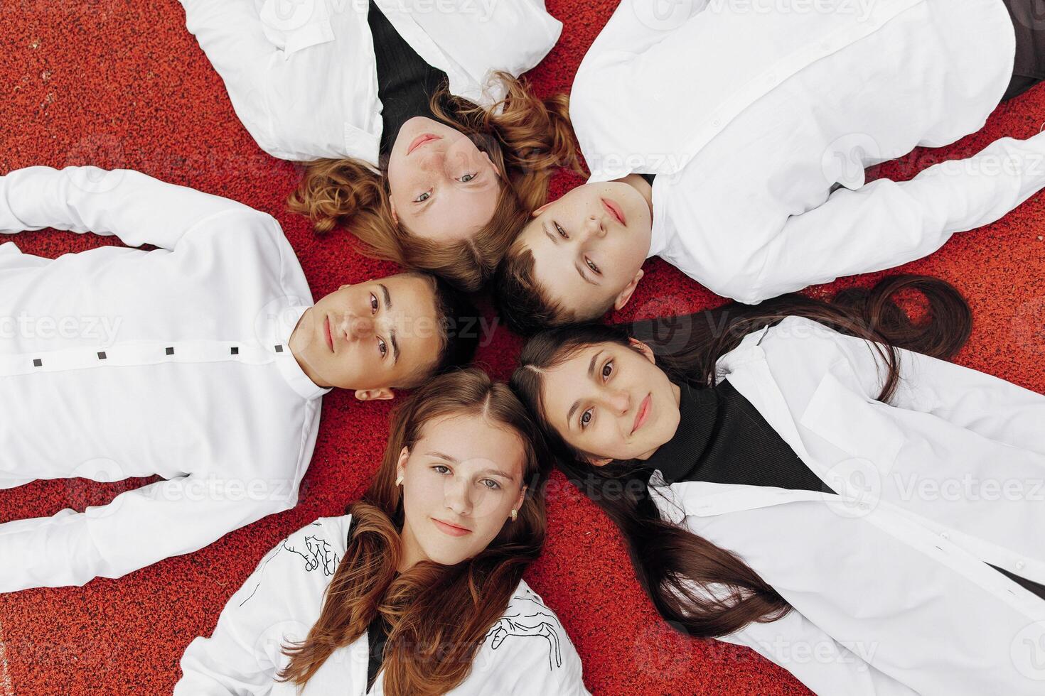 Happy friends lying together in a circle looking at the camera, holding hands and smiling while lying on a red wall... Friendship, lifestyle, togetherness, business and teamwork concepts. photo