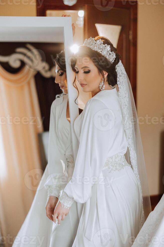 portrait of a luxurious bride in boudoir style near a mirror with reflection. Preparing the bride for the wedding in the morning in a white robe. photo